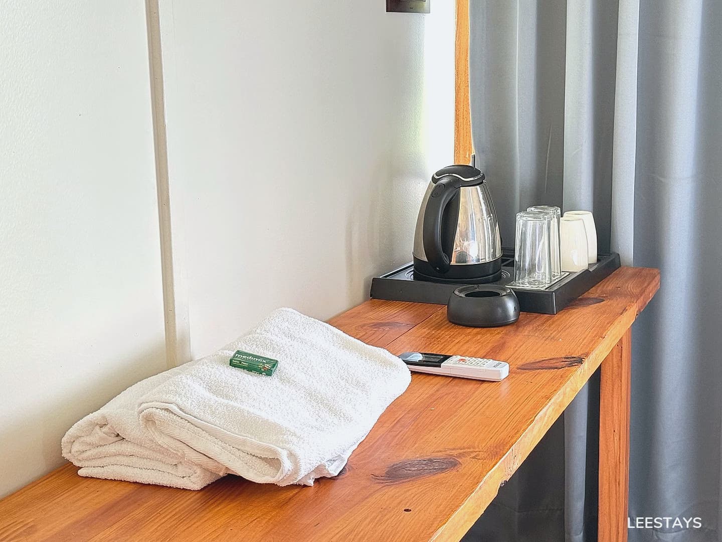 A white folded towel with a small green box on top, a remote control, an electric kettle, two clear glasses, and two white cups on a wooden table with grey curt