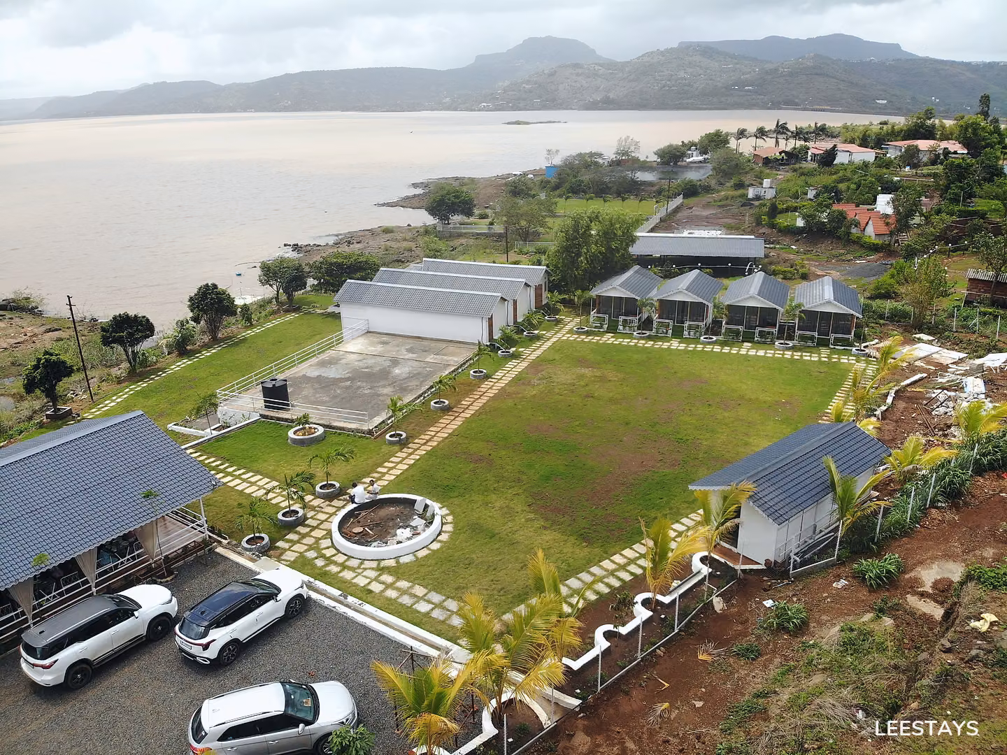 Aerial view of a resort property with multiple white cabins, a large body of water, and distant mountains under a cloudy sky.