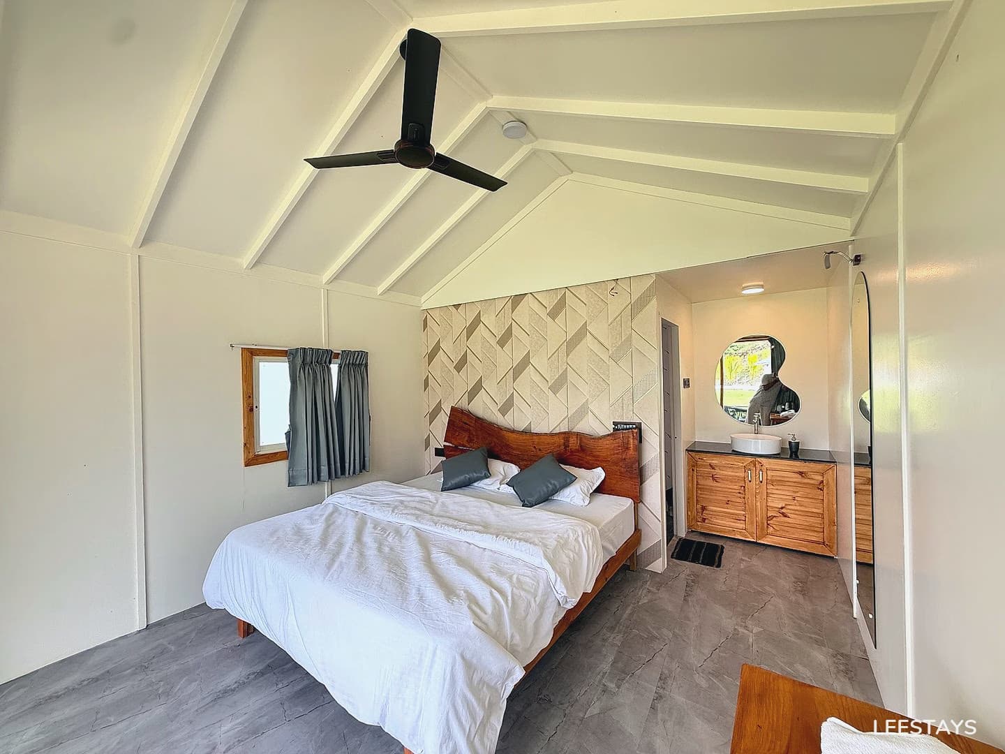 A bedroom with a large white bed, a black ceiling fan, a small window with gray curtains, and a vanity area with a sink and mirror.