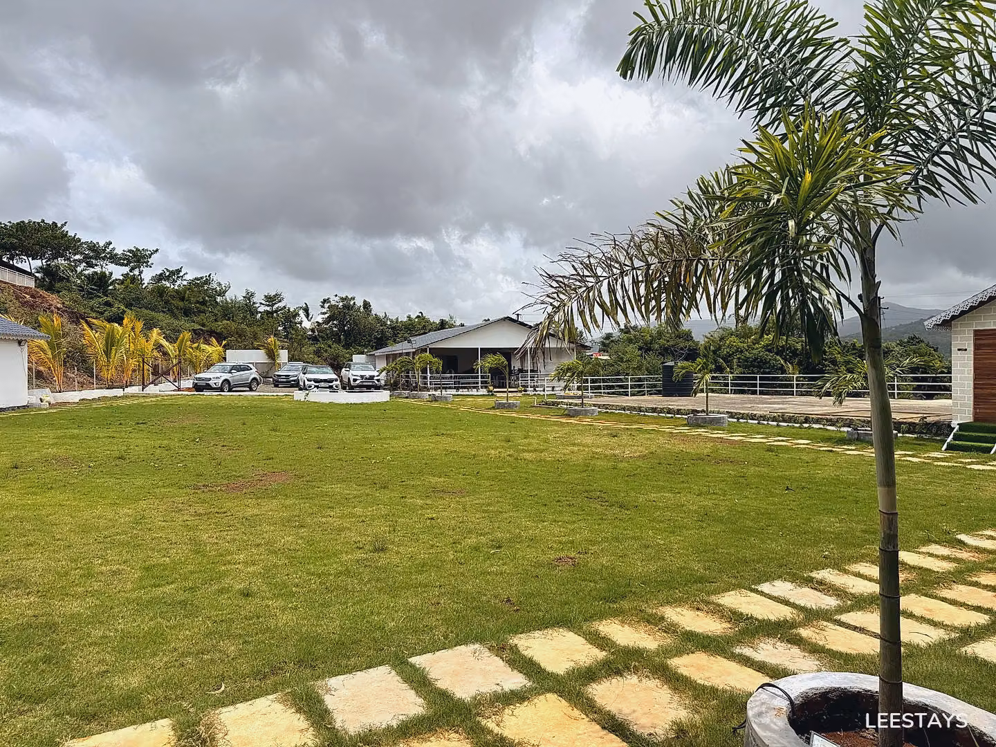 A wide green lawn with several cars parked in front of a building with a white roof and palm trees surrounding the property.