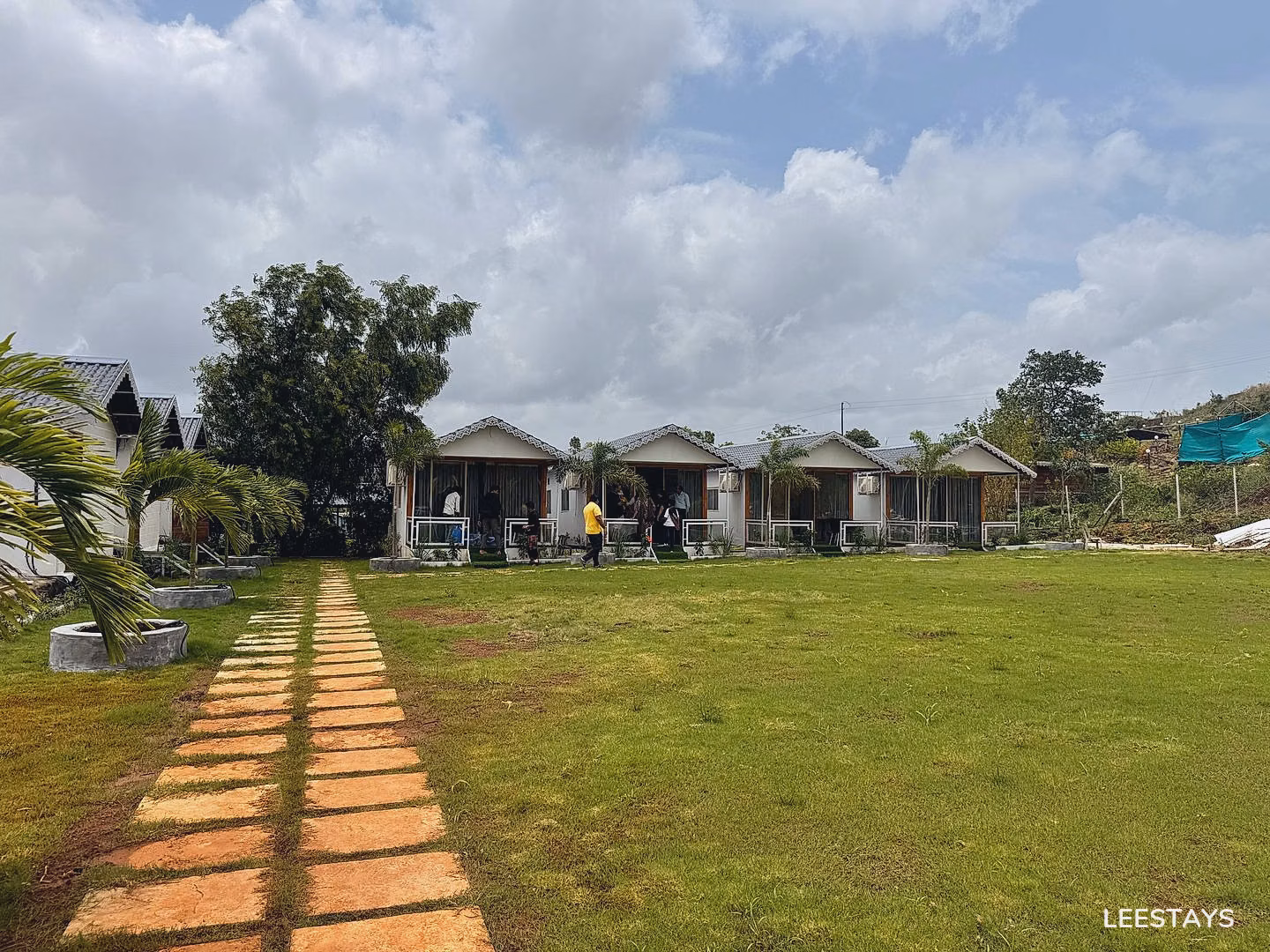 A row of small white cabins with pitched roofs and glass doors, set back from a grassy lawn. A stone pathway leads towards the cabins, flanked by green grass an