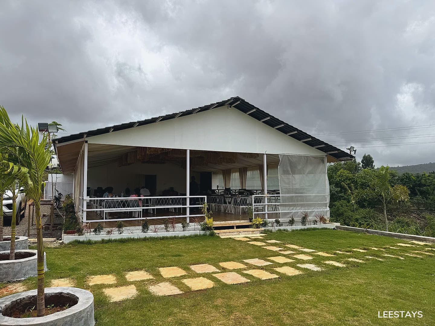 Outdoor dining area with tables and chairs under a covered patio with sheer curtains, surrounded by green grass and trees under a cloudy sky.