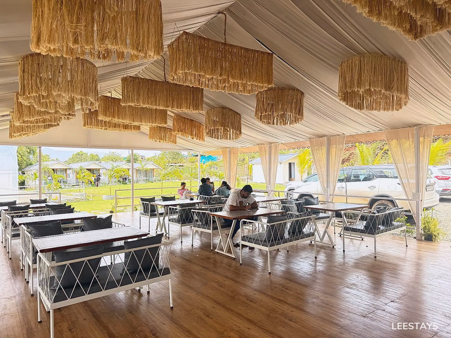 Outdoor dining area with white tables and chairs, rustic straw chandeliers hanging from a white tent ceiling, and a view of bungalows in the distance.