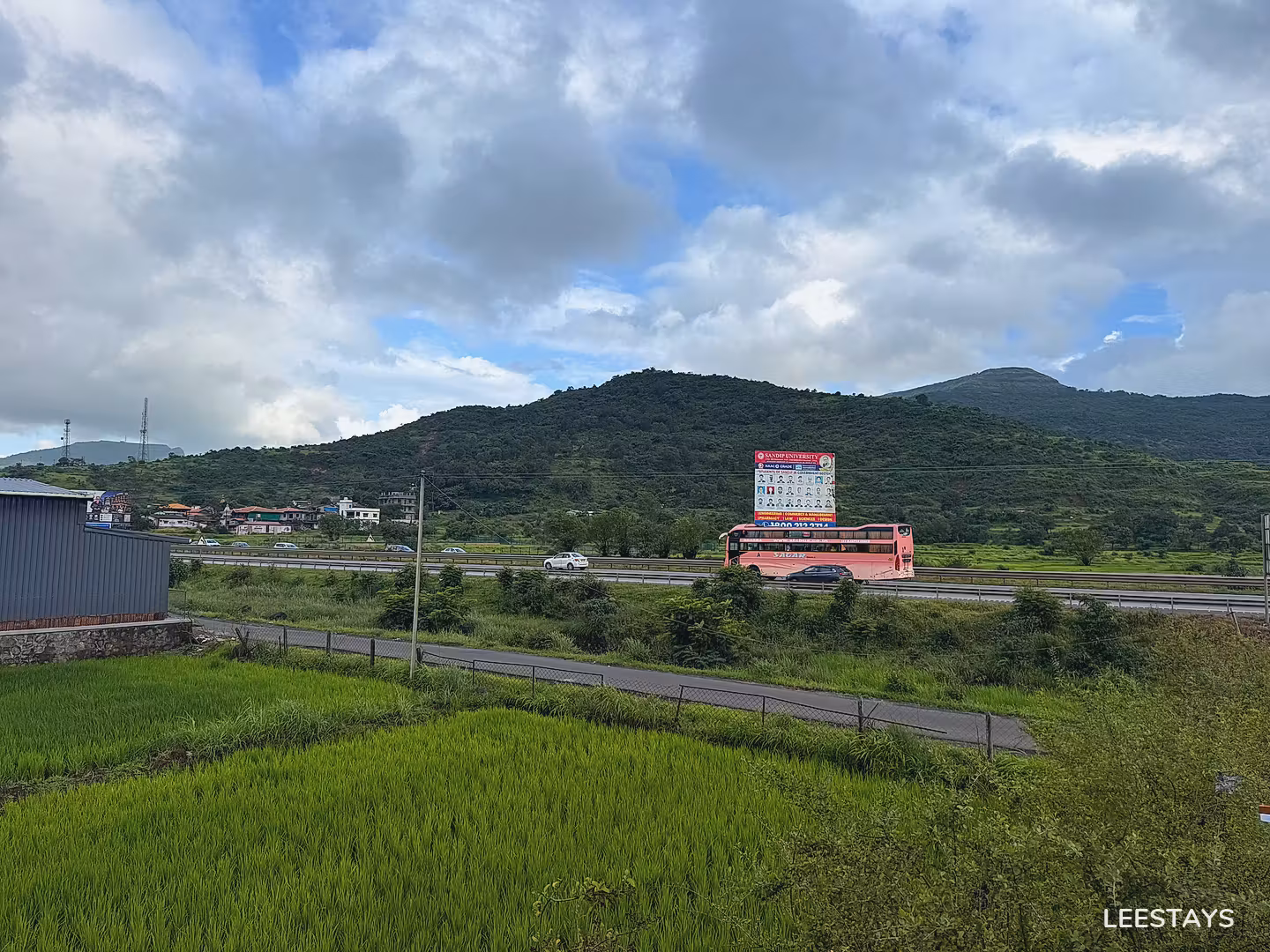Green agricultural field with a highway and wooded hills in the background under a cloudy sky.
