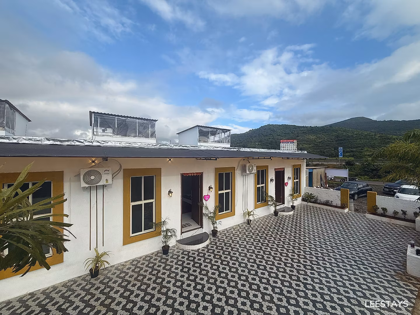 White building with yellow-trimmed windows and air conditioning units, facing a patterned paved area and a green, hilly landscape under a cloudy sky.
