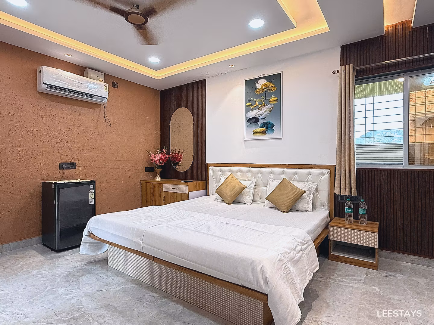 King bed with white linens, gold accent pillows, and a tufted headboard in a room with a mini-fridge and air conditioning.