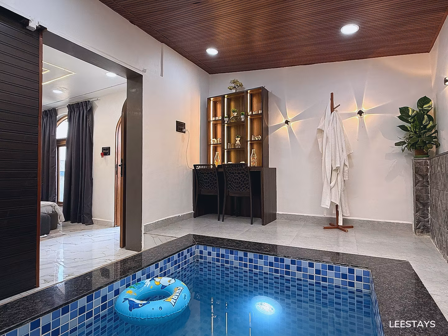 Small indoor pool with blue tiling and a child's inflatable ring, next to a desk with chairs, and a rack with robes.
