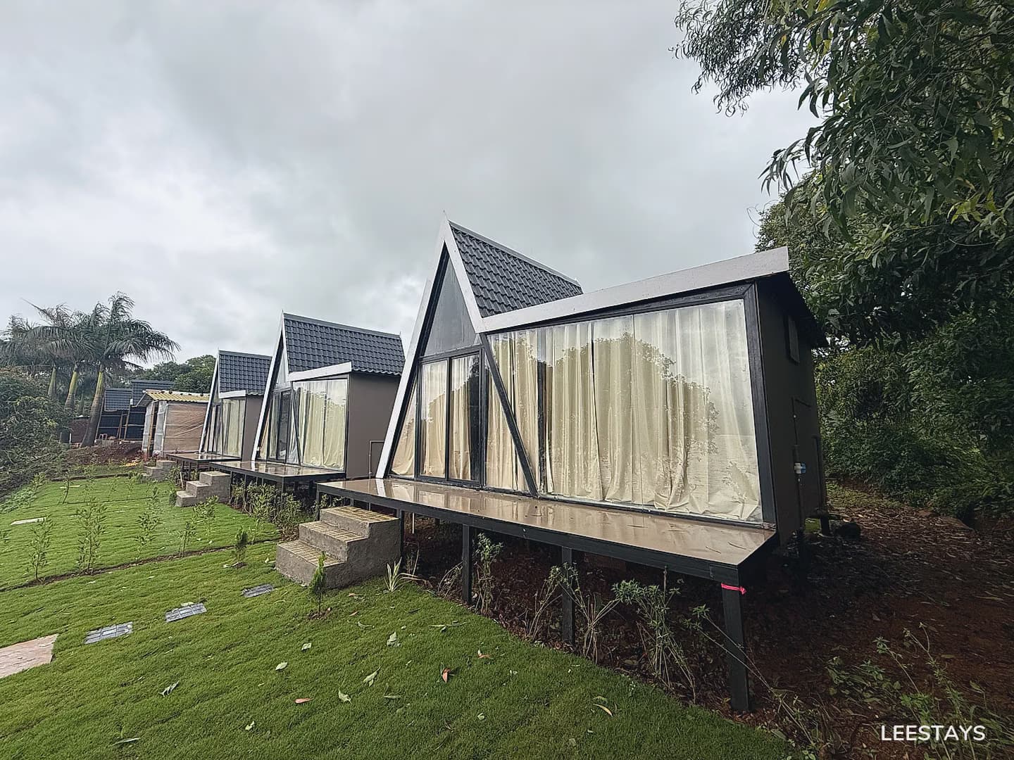 Multiple triangular prism-shaped cottages with large glass windows and peaked roofs are lined up on a sloped grassy hillside. Each cottage has a deck with stair