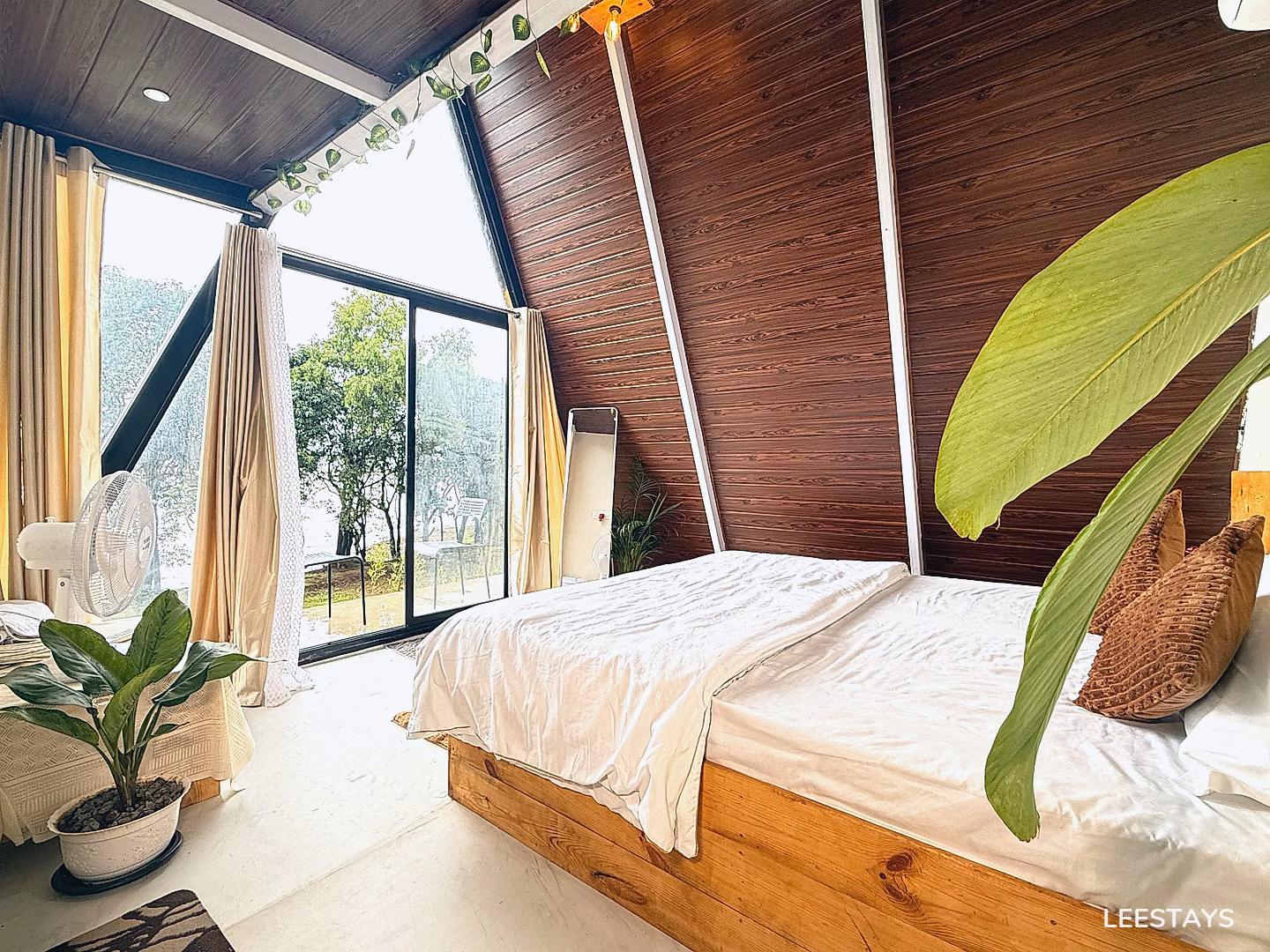 A bed with white linens is placed in a room with a sloped wooden ceiling and a large window overlooking trees and water.