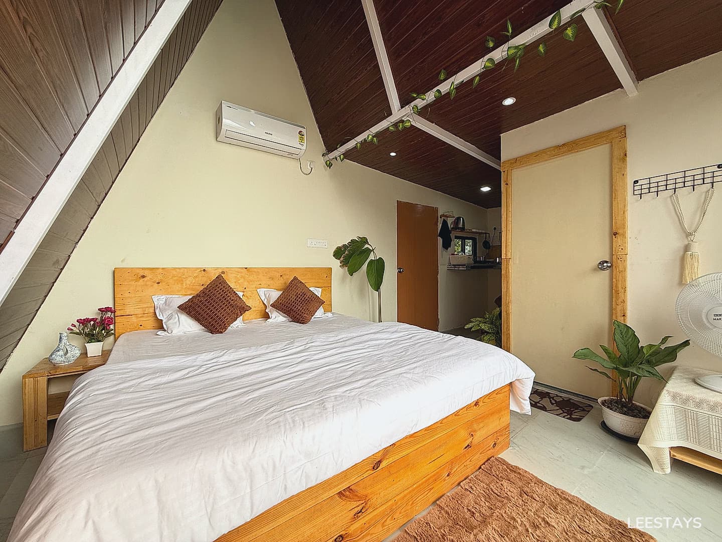 A bed with white linens and brown pillows is in a room with a slanted wooden ceiling and an air conditioning unit on the wall. Plants are placed around the room