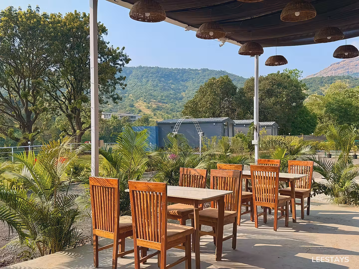Wooden tables and chairs set up outdoors with lush greenery and mountains in the background.