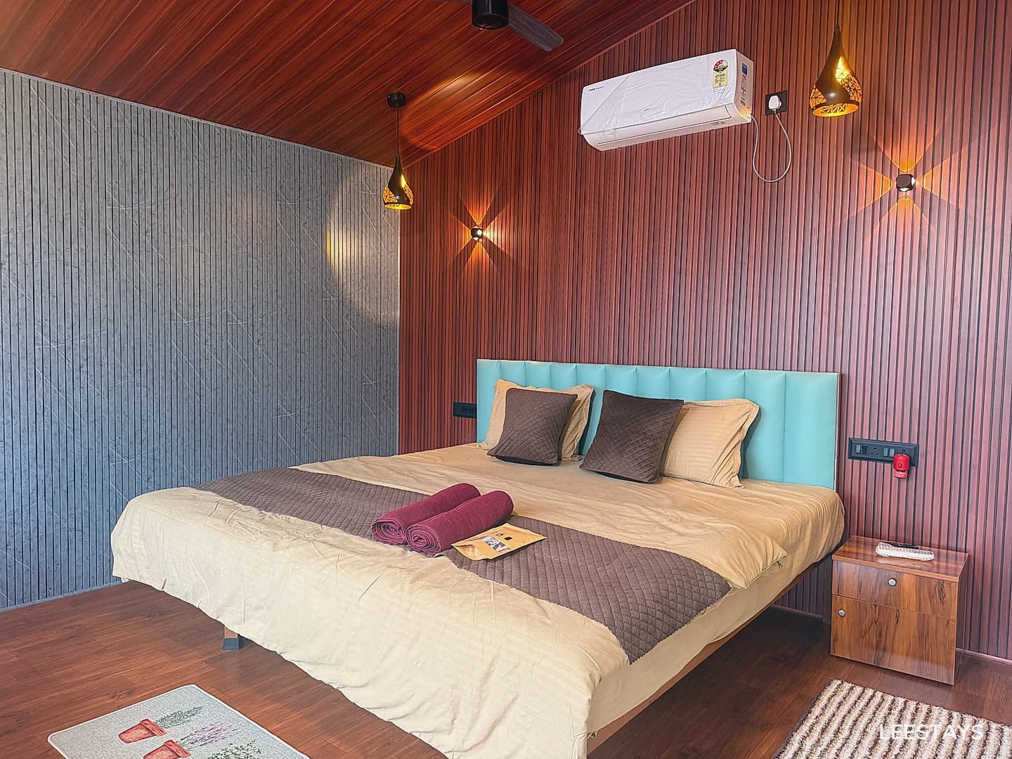 A king-size bed with a light blue headboard, tan duvet, and brown quilted throw. Two rolled towels and a pamphlet rest on the bed. The walls have wood paneling