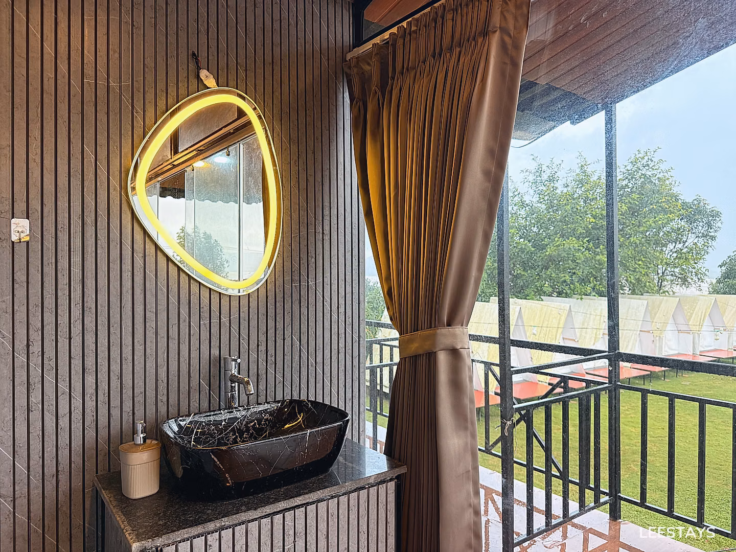 Modern bathroom vanity with a black marble sink and a uniquely shaped mirror with an illuminated border, next to a window with beige curtains showing tents and