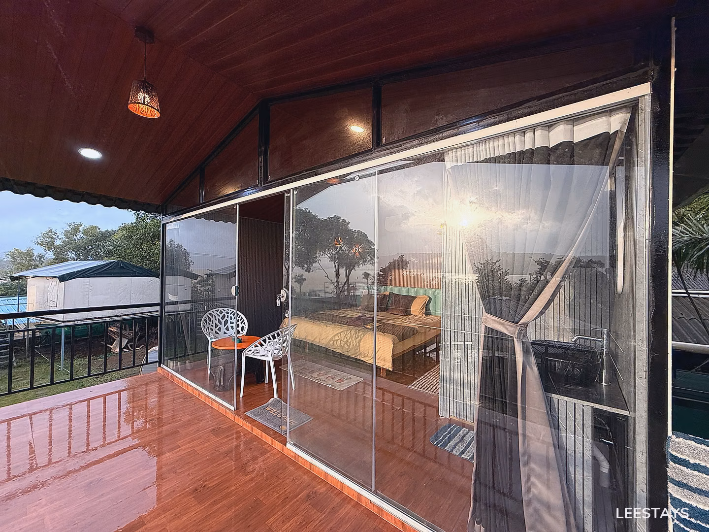 A bedroom with a large glass door showing a bed and two chairs on a balcony with a wooden floor and a view of trees and the sky.