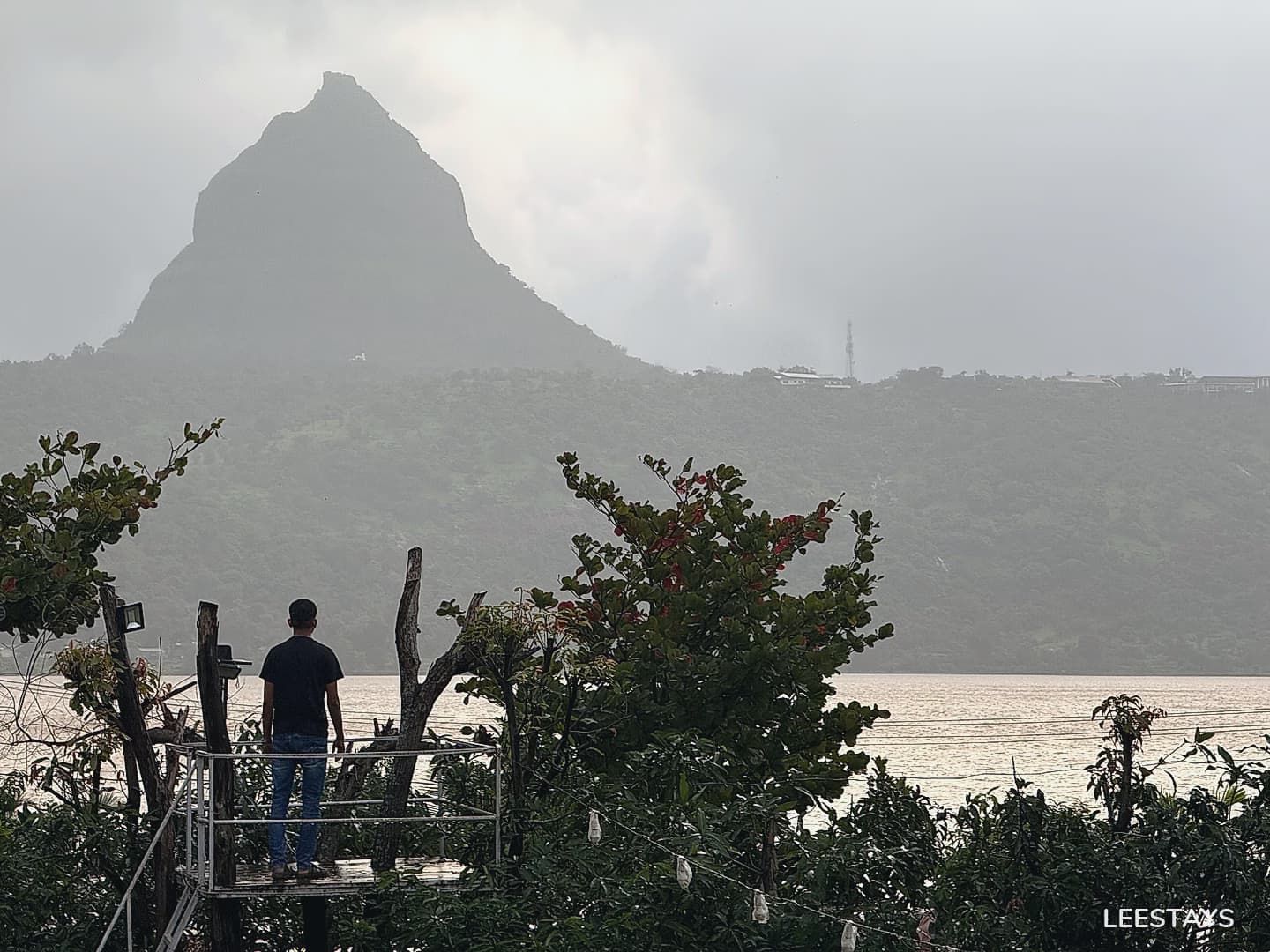 A man stands on a raised platform overlooking a calm body of water, with lush green hills and a prominent, sharp peak in the misty background.