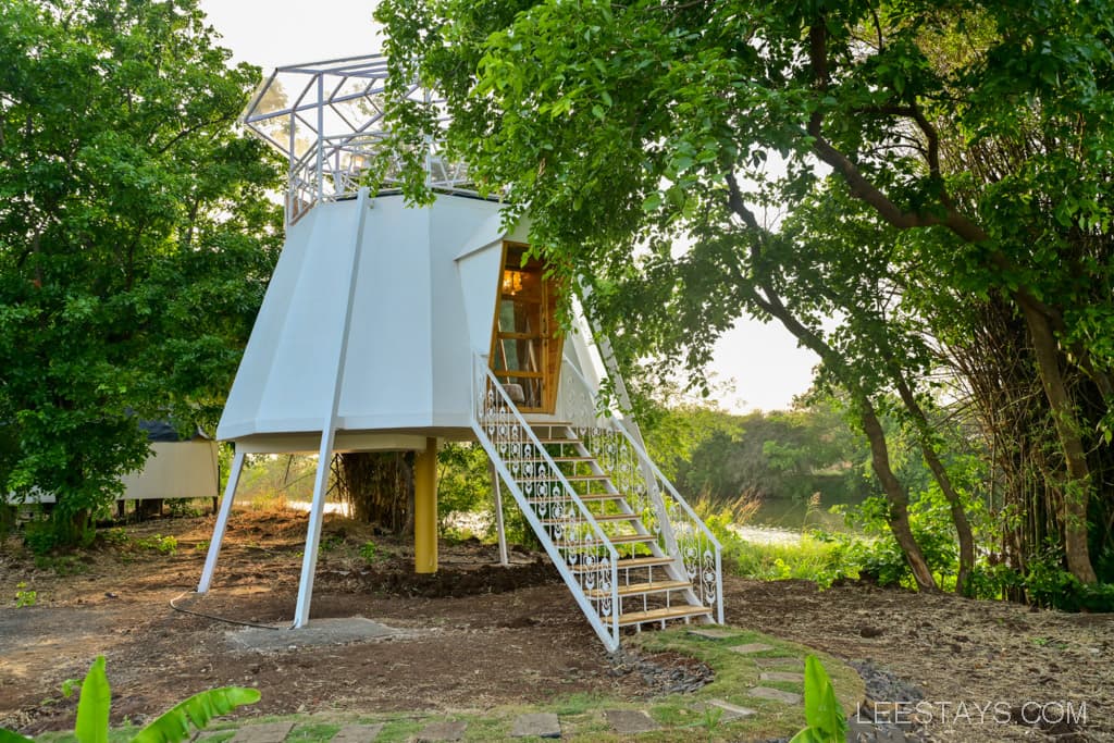 A modern treehouse with a white exterior and a staircase leading up to the entrance, surrounded by greenery.