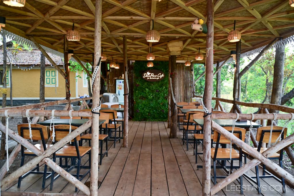 Wooden dining area featuring tables and chairs under a thatched roof with natural elements and greenery.