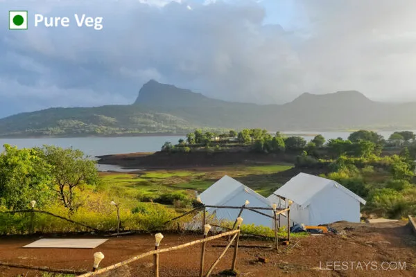 Scenic view of glamping set up on a grassy hilltop with a mountain and Pawna lake in the background under a cloudy sky