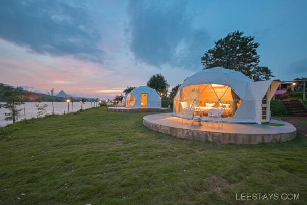 A scenic view of Dome Story Resort at Malvandi Lake, featuring two illuminated geodesic domes with a green lawn leading to the lake, captured at dusk with a colorful sky in the background.