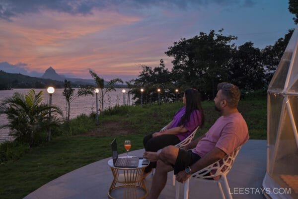 A couple sitting outside enjoying the sunset view over Malvandi Lake at Dome Story Resort, with twinkling lights and lush greenery around them.