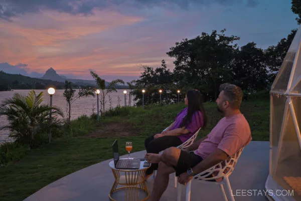 A couple sitting outside enjoying the sunset view over Malvandi Lake at Dome Story Resort, with twinkling lights and lush greenery around them.