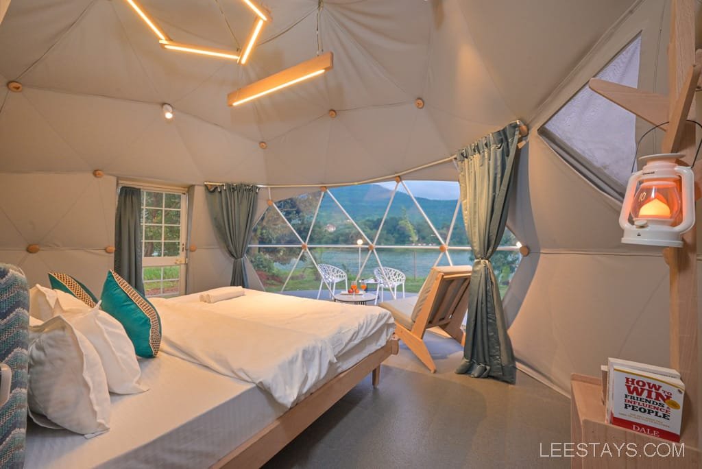 Interior of a dome room at Dome Story Resort, Malvandi Lake, featuring a double bed with pillows, a window overlooking the lake, geometric lighting, and a small table with chairs outside.