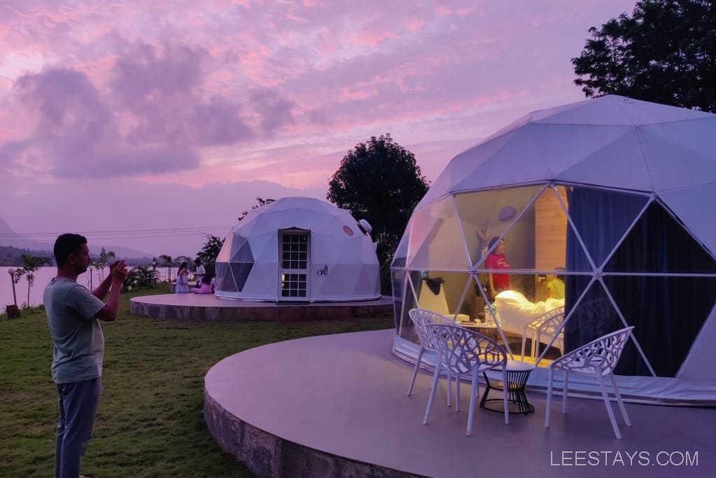 Dome-shaped cabins at Dome Story Resort by Malvandi Lake during sunset, with a person taking photos in the foreground and the interior of one cabin visible.