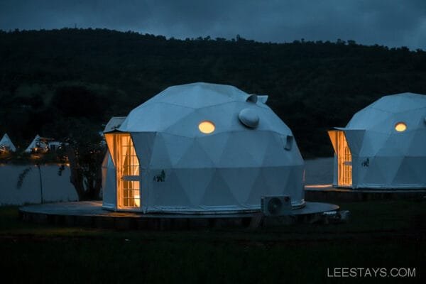 Geodesic domes lit from within at Dome Story Resort, situated by Malvandi Lake, against a backdrop of hills at dusk.