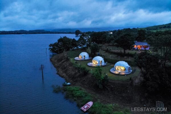 Aerial view of Dome Story Resort by Malvandi Lake, featuring several illuminated dome structures along the waterfront surrounded by trees, with a lake and cloudy sky in the background at dusk.