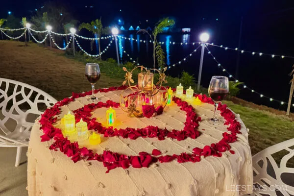 Romantic outdoor table setting at Dome Story Resort, Malvandi Lake, featuring rose petals, glowing lanterns, and glasses of red wine, with a view of the lake and string lights in the background.