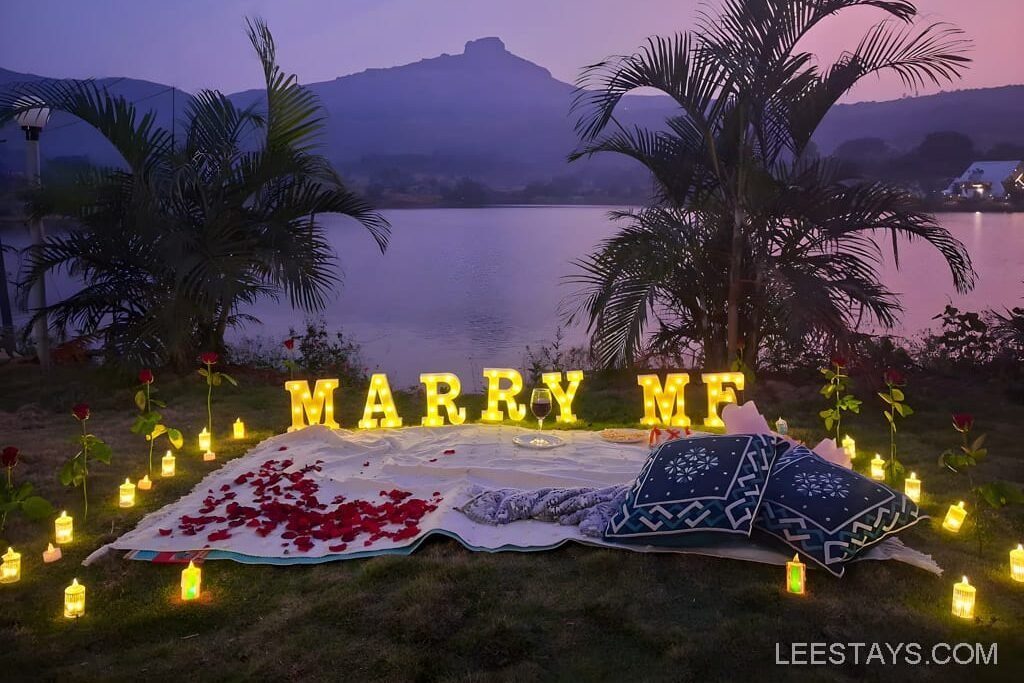 A romantic lakeside setup at Dome Story Resort, Malvandi Lake, featuring illuminated 'Marry Me' letters, surrounded by candles, rose petals, and cushions on a blanket at dusk.