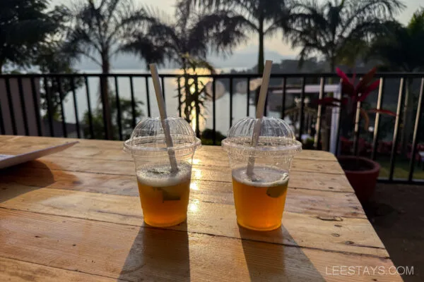 Two beverages on a wooden table with a view of Pawna Lake's serene glamping site at sunset, surrounded by palm trees and lush greenery.