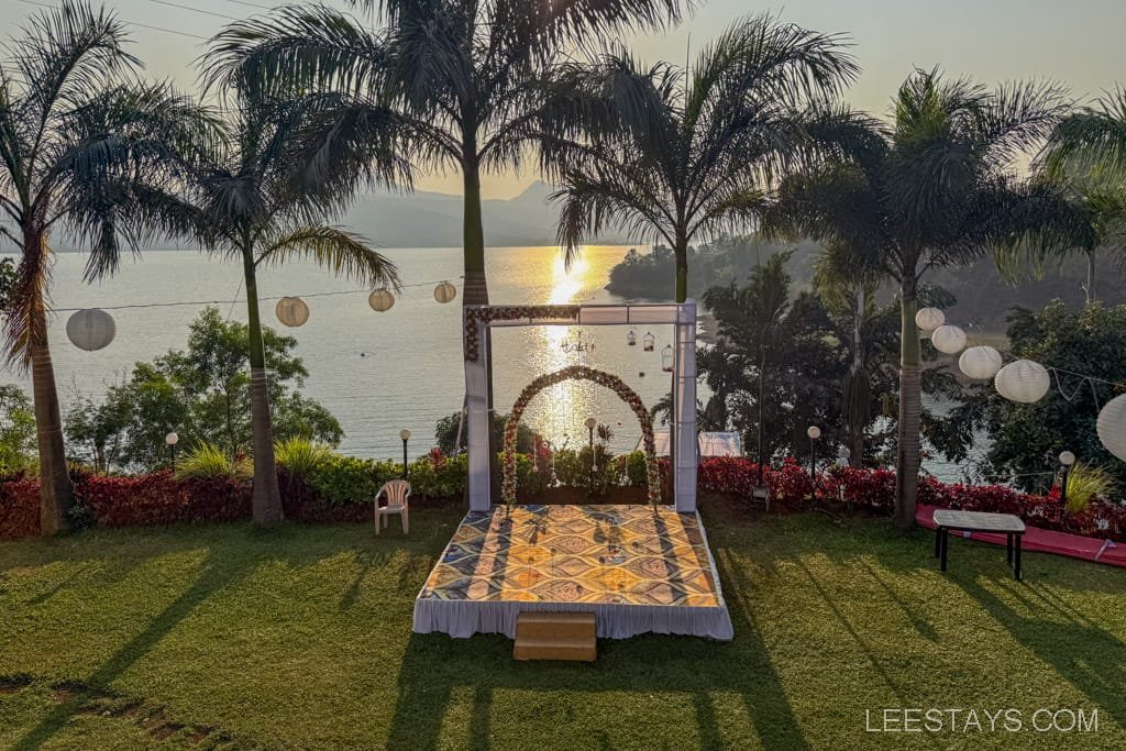 Scenic glamping spot at Pawna Lake with a decorated platform, lush greenery, palm trees, and hanging lanterns overlooking the serene water at sunset.