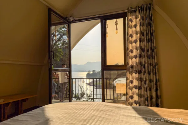 Cozy interior setup for glamping with a view of Pawna Lake through an open glass door, featuring comfortable bedding and stylish curtains.