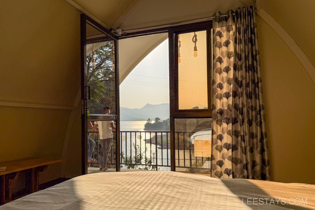 Cozy interior setup for glamping with a view of Pawna Lake through an open glass door, featuring comfortable bedding and stylish curtains.