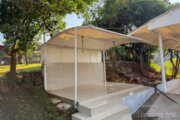 Outdoor covered area with white tiles and a roof, surrounded by greenery at Stay, Pawna; featuring a serene natural setting. LEESTAYS.COM.