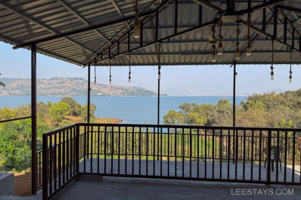 A scenic view of Pawna Lake from a sheltered balcony, featuring a metal roof and hanging lights, surrounded by lush greenery, ideal for a relaxing stay.