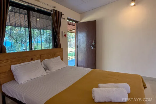 A cozy bedroom at Stay in Pawna featuring a wooden bed with white sheets and towels, a large window with curtains, and an open door leading outside.