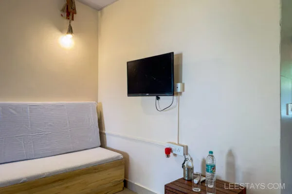 A cozy room at Stay, Pawna, featuring a wall-mounted TV, a light fixture, a cushioned bench, and a wooden side table with bottled water and glassware.