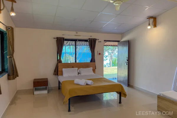 A cozy, well-lit room at Stay, Pawna featuring a bed with a mustard-colored blanket, a small table, and an open door leading to a grassy area outside.