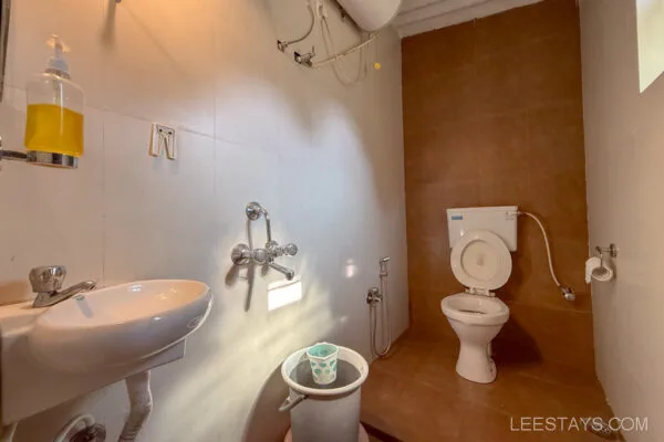 Bathroom at Stay in Pawna with white tiles, a toilet, sink, bucket, and soap dispenser, providing basic amenities for guests.