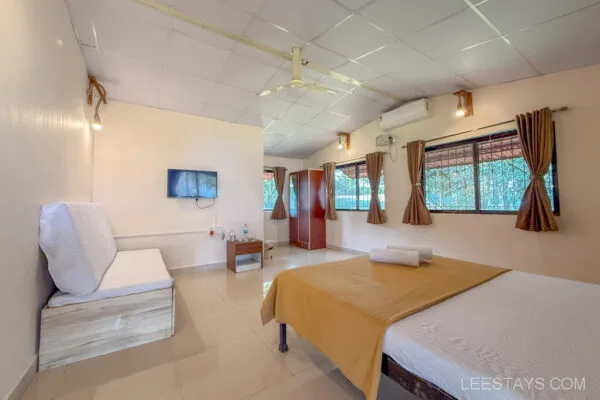 Interior of a cozy Stay in Pawna featuring a bed with a mustard blanket, a sofa, TV mounted on the wall, curtains, and ample natural light through windows.