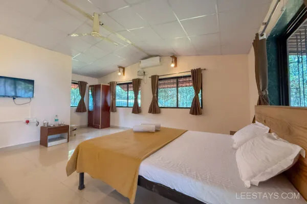 Cozy bedroom at Stay, Pawna, featuring a neatly arranged bed with mustard bedding, air conditioning, a wall-mounted TV, and large windows with brown curtains.