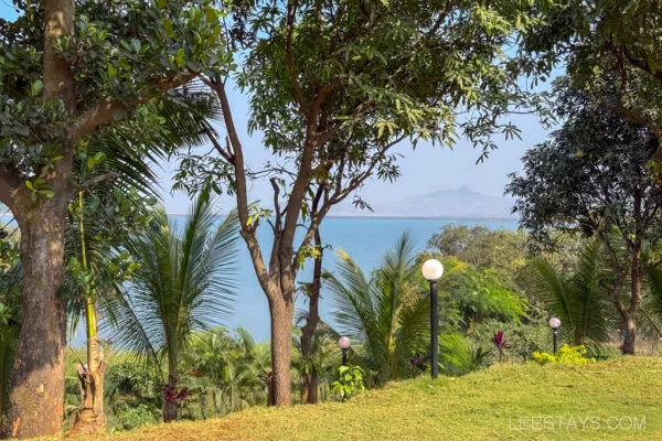 Scenic view of a lush green landscape with tall trees framing Pawna Lake, offering a tranquil stay at Lee Stays, Pawna, under a clear blue sky.