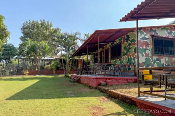 A camouflaged bungalow with a covered porch and seating area, surrounded by trees and a manicured lawn, named Stay, Pawna.