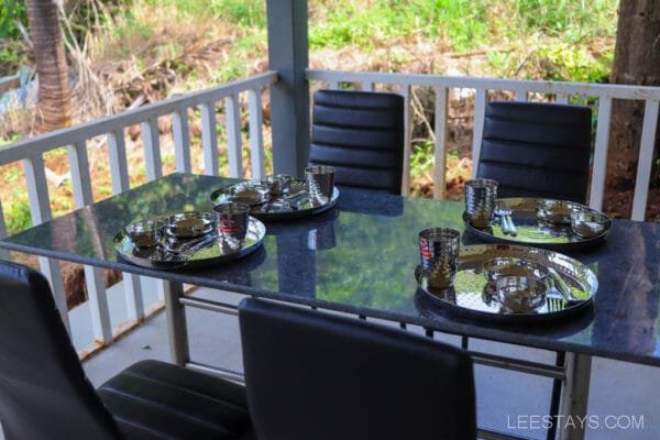 Dining table set outdoors at a resort in Lonavala, featuring metallic plates, glasses, and cutlery, surrounded by lush greenery.