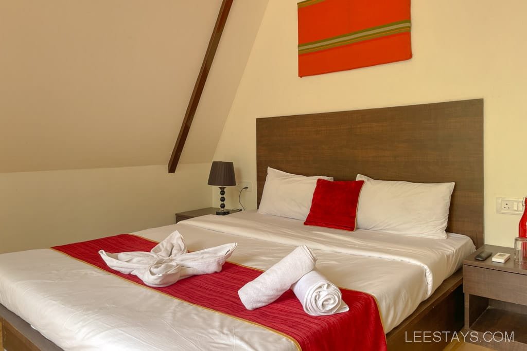 Cozy resort room in Lonavala featuring a neatly made bed with red and white accents, a bedside table, and stylish decor in a serene setting.