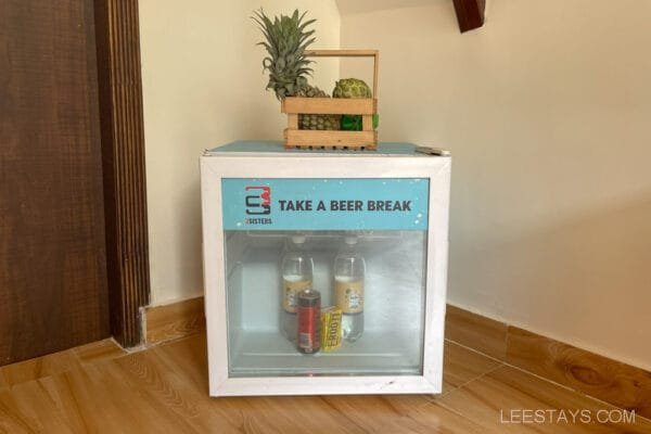 A small fridge labeled 'Take a Beer Break' in a resort in Lonavala, containing drinks and topped with a wooden basket of fresh fruits.