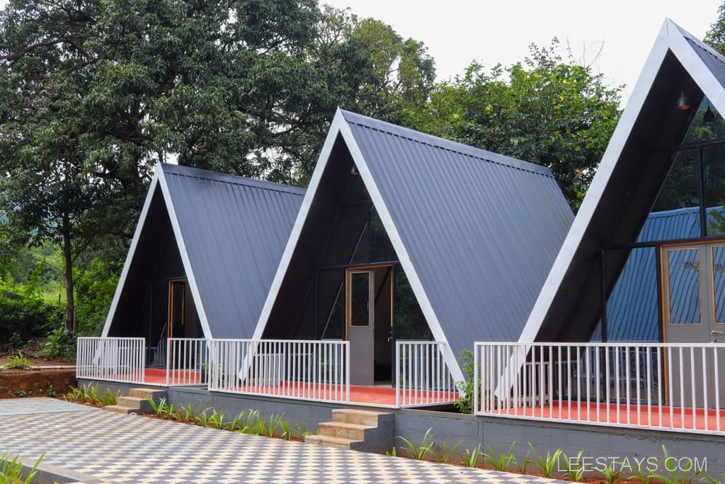 Modern resort cabins in Lonavala featuring triangular roofs, surrounded by lush greenery, with a patterned walkway leading to the entrance.