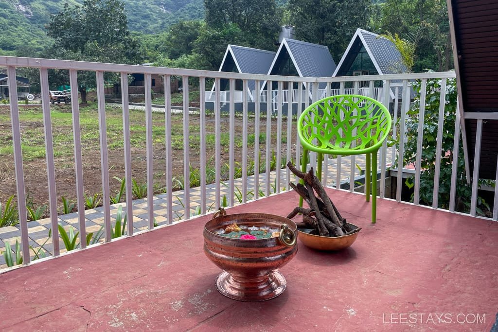 A cozy balcony at a resort in Lonavala featuring a bright green chair, a decorative bowl with flower petals, and a bundle of firewood, with scenic hills in the background.